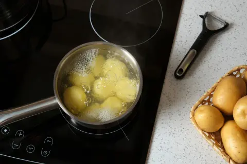Ziemniaki gotują się w garnku na płycie indukcyjnej. Obok leży obieraczka i koszyk z surowymi ziemniakami. To początek, jak ugotować ziemniaki.