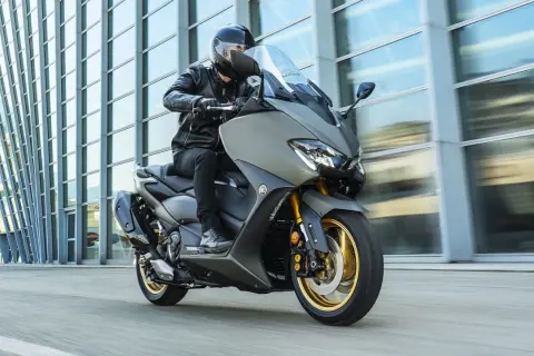 Hombre en moto clásica moderna, con chaqueta de cuero, circulando frente a un edificio de cristal.