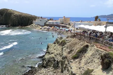 Gente disfrutando en un restaurante de Cabo de Gata, con vistas a una playa concurrida y el mar azul.