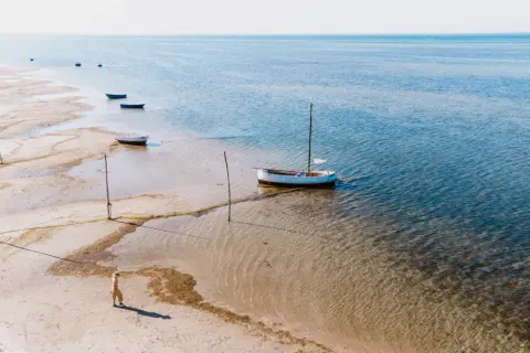 Spokojna plaża w Kuźnicy, kilka łodzi na płytkiej wodzie, jedna z masztem. Osoba w słomkowym kapeluszu spaceruje brzegiem.