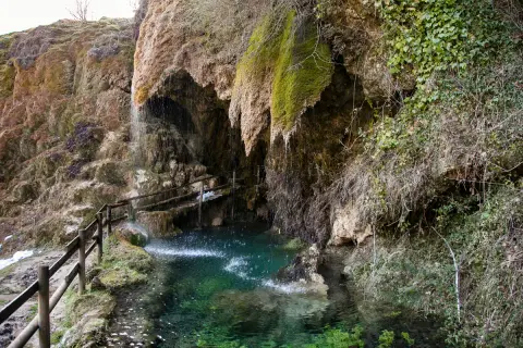Cascata che scende tra rocce muschiose, formando una pozza d'acqua cristallina. Un sentiero con staccionata in legno conduce alle grotte di Labante.