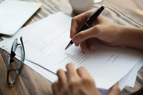 Dłoń z długopisem nad dokumentem, podpisując umowę. W tle okulary i tablet. Kluczowe jest porozumienie stron.