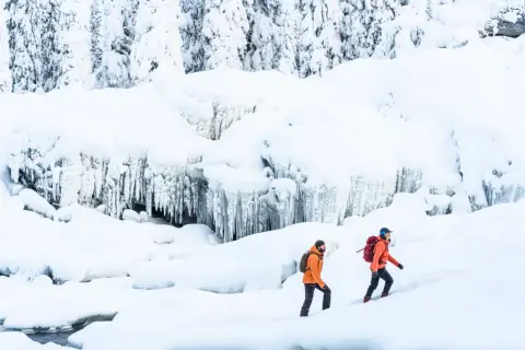 Två personer med ryggsäckar vandrar på vintern i ett snöigt landskap med istappar.