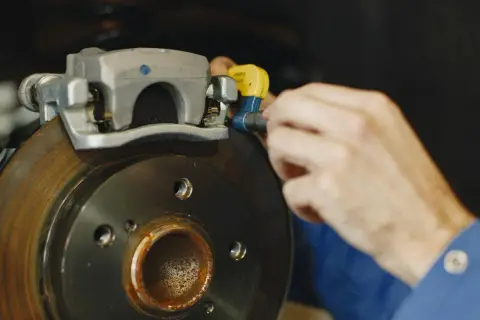 Mechanik dokonuje wymiany linki hamulca ręcznego, pracując przy zacisku i tarczy hamulcowej.