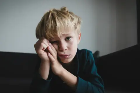 Chłopiec z blond włosami, zamyślony, opiera głowę na dłoniach. Jego spojrzenie sugeruje trudności, być może związane z zespołem Aspergera.
