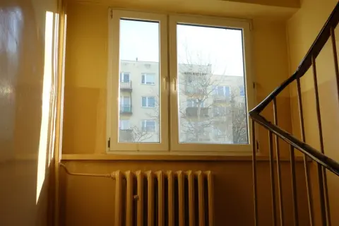 Słoneczne promienie padają na błyszczącą ścianę w klatce schodowej. Widać okno z widokiem na blok i kaloryfer.
