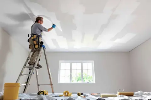 Un peintre sur une échelle applique une toile de rénovation plafond. Des rouleaux et un seau sont prêts.