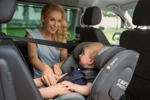 Lächelnde Mutter sichert ihr Baby im Kindersitz vorne im Auto.
