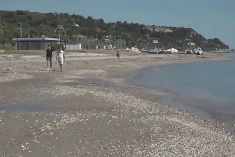 Passeggiata sulla spiaggia di Marina di Campofilone, con due uomini che camminano e barche ormeggiate sullo sfondo.