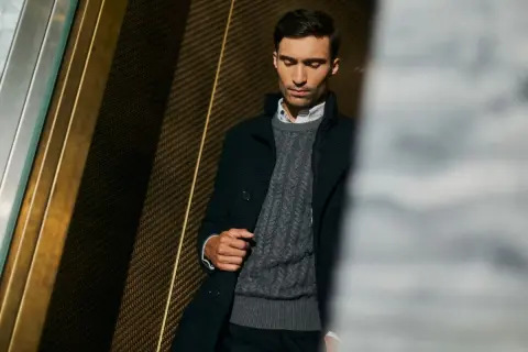Elegancki mężczyzna w szarym swetrze męskim z warkoczowym splotem, pod ciemną marynarką. Stylowa inspiracja, jak nosić sweter męski.
