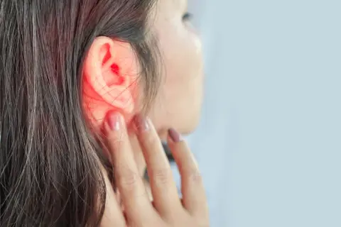 Une femme se touche l'oreille, une zone rouge vif indiquant une douleur ou une inflammation. Un point noir est visible dans l'oreille.