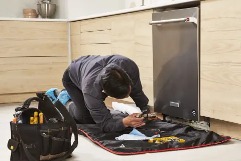A technician kneels to replace a dishwasher, surrounded by tools and a protective mat.