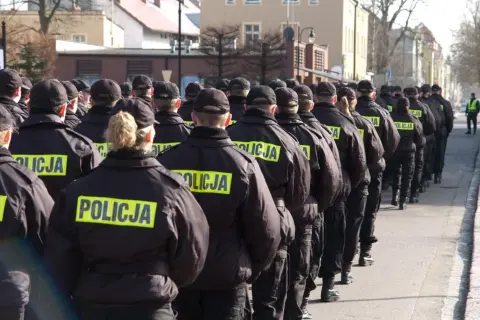 Policjanci w czarnych kurtkach z napisem "POLICJA" stoją w szeregu, gotowi do akcji. Widać, jak pracuje policja zmiany, przygotowując się do służby.