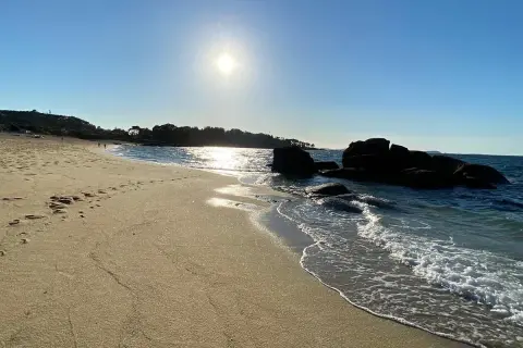 La **playa de Tulla** bajo un sol radiante. Huellas en la arena dorada y el mar azul acarician las rocas oscuras.