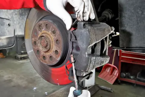 Mechanik w białych rękawiczkach dokonuje wymiany klocków hamulcowych, używając śrubokręta do podważenia starego klocka.