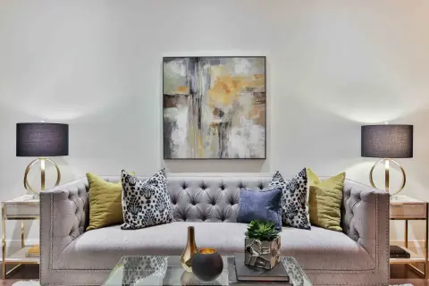 Nowoczesna szara sofa z pikowanymi guzikami, ozdobiona poduszkami i abstrakcyjnym obrazem. To idealne miejsce na relaks, tworzące przytulną atmosferę.