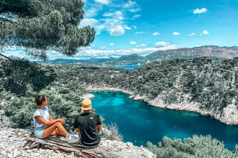 Deux personnes assises sur un tronc d'arbre contemplent une crique aux eaux turquoise, entourée de falaises rocheuses et de pins.