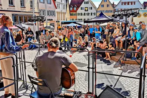 Ein Musiker spielt Gitarre, während eine Sängerin auf einer Bühne steht. Viele Menschen sitzen und stehen davor, um die **Veranstaltung weil der Stadt** zu genießen.