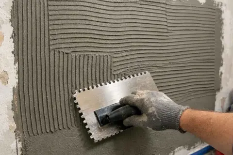 Main tenant, une truelle dentée pour étaler la colle carrelage préparée sur un mur. Les sillons créés par la truelle sont visibles.
