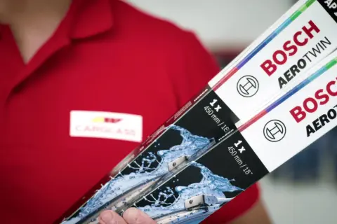 Person i röd pikétröja med Carglass-logga håller upp två paket med Bosch Aerotwin torkarblad.