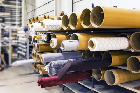 Różne rodzaje materiałów na rolkach, gotowe do użycia w produkcji. Od jasnych wzorzystych tkanin po ciemne, jednolite materiały.