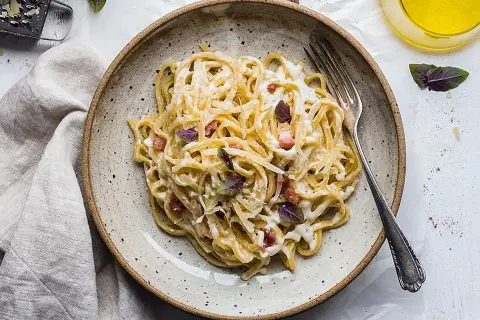 En tallrik med krämig, klassisk carbonara, toppad med basilika och parmesan. En gaffel ligger redo att avnjutas.