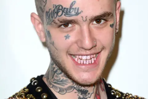 Uśmiechnięty Lil Peep z tatuażami na twarzy i szyi, błyszczącymi różowymi zębami. Pytanie czy lil peep żyje, pozostaje w sercach fanów.