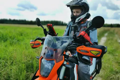 Motocyklista w kasku i kurtce przygotowuje się do podróży motocyklem po Europie. W tle zielone pole i niebo.