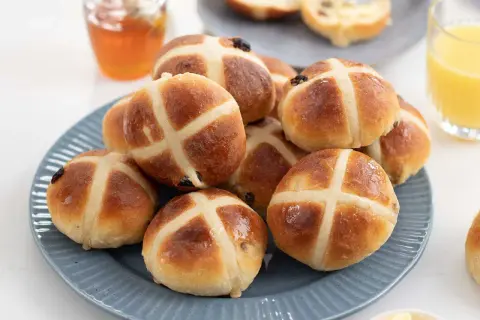 En hög med gyllene, nybakade **hot cross buns** på en blå tallrik, redo att avnjutas med honung och apelsinjuice.