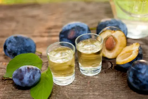 Dwa kieliszki ze złotym płynem, otoczone dojrzałymi śliwkami i liśćmi. To tradycyjny albański alkohol, idealny na letnie wieczory.
