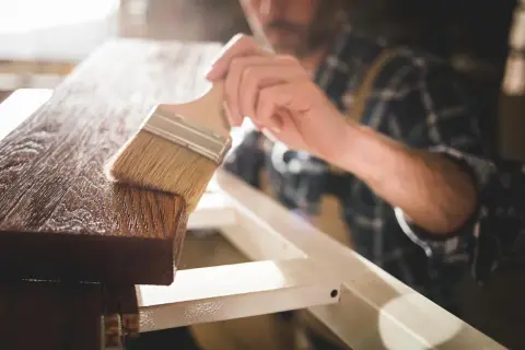 Un falegname applica una finitura protettiva su un pezzo di legno. Dopo l'impregnante, cosa si mette? Una vernice trasparente o una cera per esaltare la venatura.