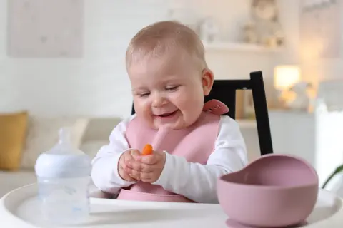 Uśmiechnięte niemowlę w różowym śliniaku trzyma marchewkę, rozpoczynając przygodę z żywieniem niemowląt. Obok butelka i miseczka.