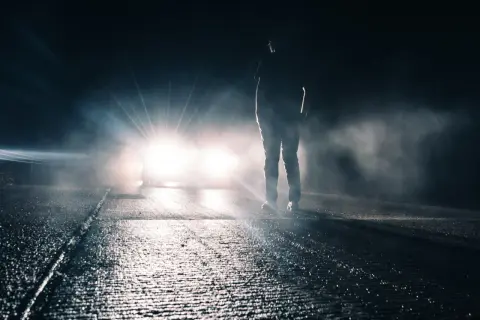 Silhouette d'une personne sur une route mouillée, éclairée par un appel de phare éblouissant dans la nuit brumeuse.