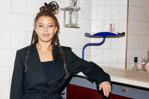 Matilda Jackson, med flätor uppsatta i en knut, lutar sig mot en bardisk med flaskor i bakgrunden.