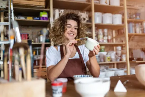 Uśmiechnięta artystka maluje ceramikę w swojej pracowni. To miejsce, gdzie powstaje i sprzedawana jest produkcja i sprzedaż rękodzieła pkd.