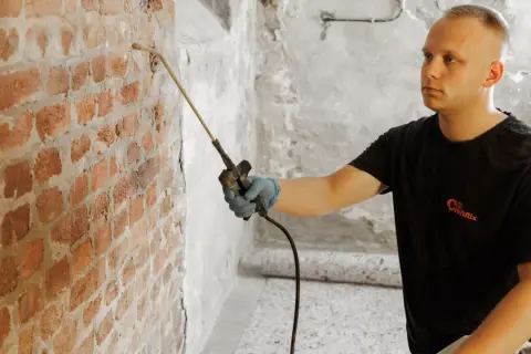 Mann sprüht mit Gerät gegen Feuchtigkeit im Keller. Ziegelwand wird behandelt.
