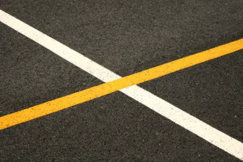 Nowe malowanie pasów drogowych: biała i żółta linia przecinają się na ciemnym asfalcie, tworząc geometryczny wzór.