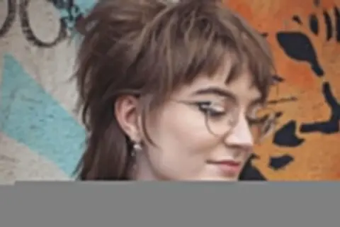 Dziewczyna w okularach z charakterystyczną, warstwową fryzurą mullet, pozuje na tle kolorowego graffiti.