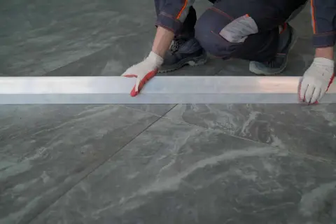 Man con guantes blancos y rojos coloca una regla metálica sobre azulejos rectificados, evitando problemas de desnivel.