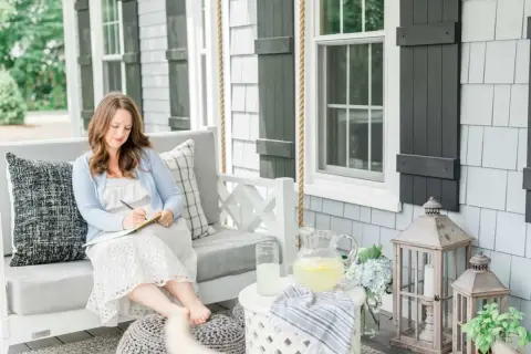 A woman embodies the coastal grandma aesthetic, writing in a notebook on a porch swing, with lemonade and hydrangeas nearby.
