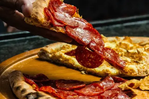 Kawałek pizzy pepperoni unoszony nad całą pizzą na drewnianej desce.