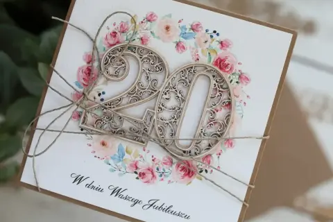 Elegancka kartka z napisem "W dniu Waszego Jubileuszu" i ażurowymi cyframi "20" na tle kwiatowej girlandy. Idealna na 20 rocznicę ślubu życzenia.