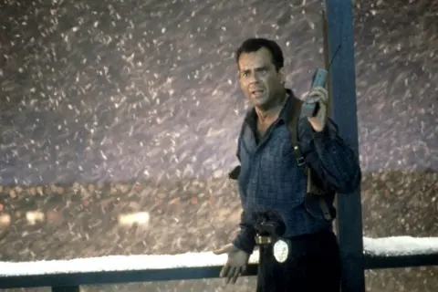John McClane, med en gammal mobiltelefon i handen, står i snöstormen. Han är en del av rollistan i Die Hard 2.