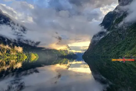 Majestatyczne fiordy norweskie, gdzie słońce przebija się przez chmury, a kajakarz przemierza spokojne wody.