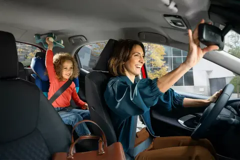 Glad mamma justerar backspegeln i sin Toyota, med barnet i bakgrunden som leker med en leksak. Med Toyota garanti kan du känna dig trygg.