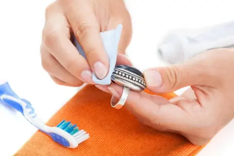 Manos limpiando un anillo de plata con un paño azul. Se ve un cepillo de dientes y una pasta de dientes, indicando cómo se limpia la plata en casa.