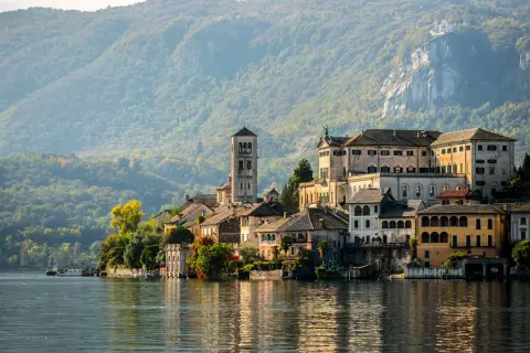 A picturesque Italian village with colorful buildings and a tall bell tower sits on the edge of a calm lake, with tree-covered mountains in the background.