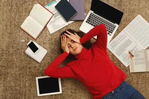 Zmęczona studentka leży na dywanie otoczona książkami, laptopem i telefonem.