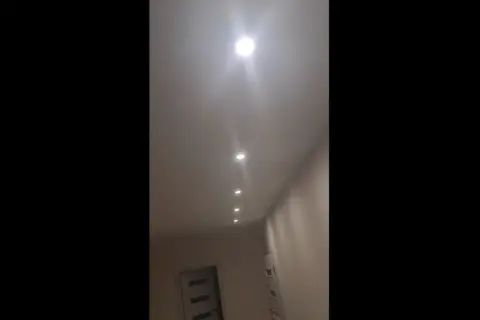 W korytarzu domu widać rząd wbudowanych w sufit lamp. Jedna z nich, najbliżej widza, świeci najjaśniej, tworząc efekt migającego światła.