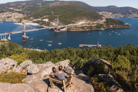 Pareja sentada en un mirador, contemplando el impresionante puente de Rande sobre el mar, con bateas de mejillones y un pueblo al fondo.